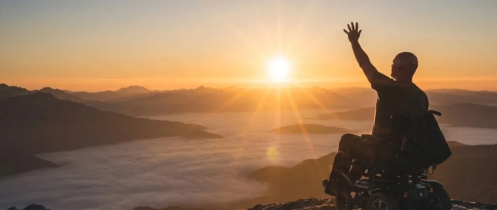 Philippe Bocquier en fauteuil roulant au sommet d'une montagne, bras levé face au lever de soleil, mer de nuages en contrebas